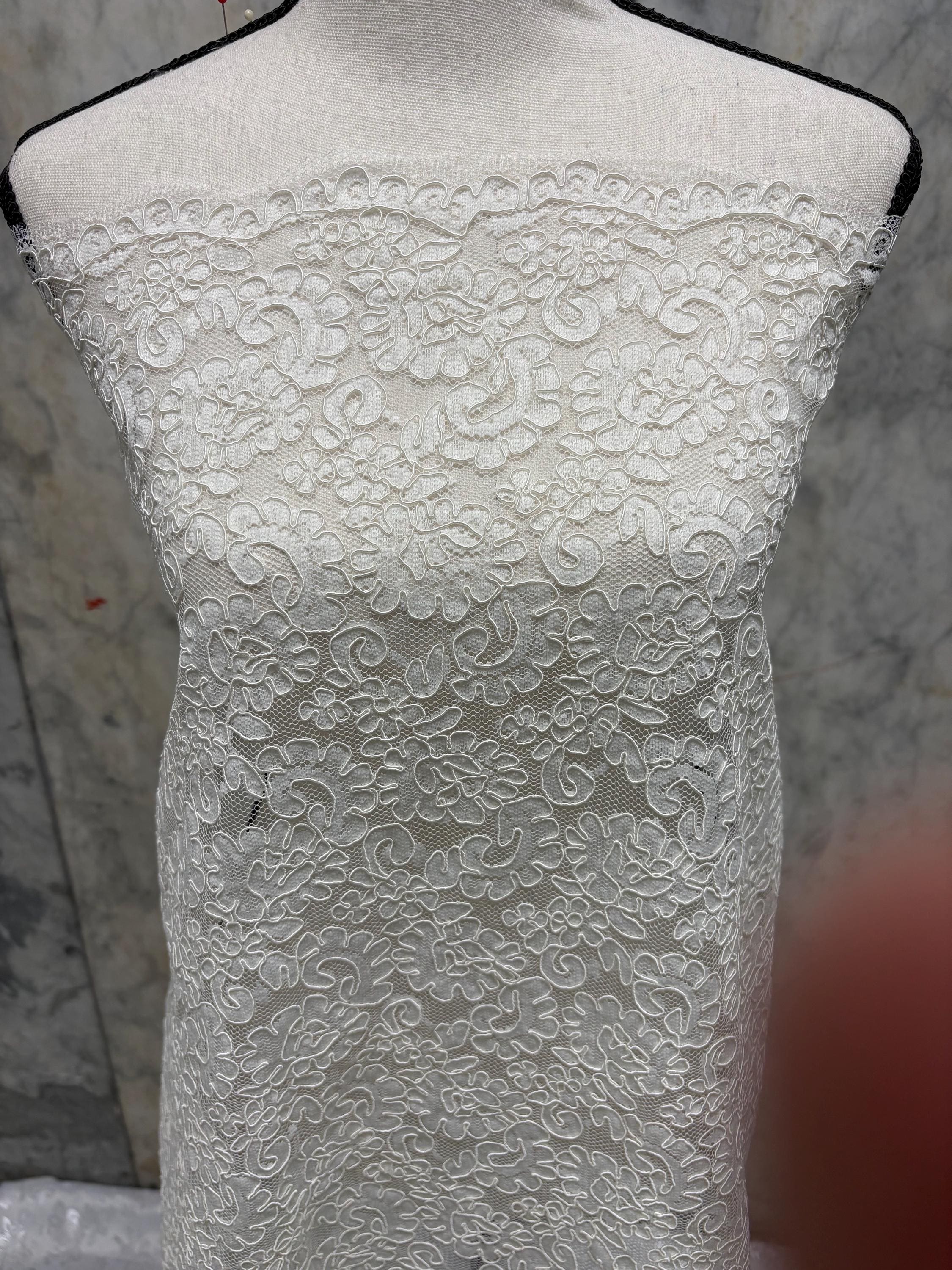 May include: Ivory lace fabric displayed on a mannequin. The fabric features a detailed floral pattern with a scalloped edge. The lace is densely woven, creating a textured surface. The fabric is draped, showcasing its delicate design and potential for apparel or decorative use