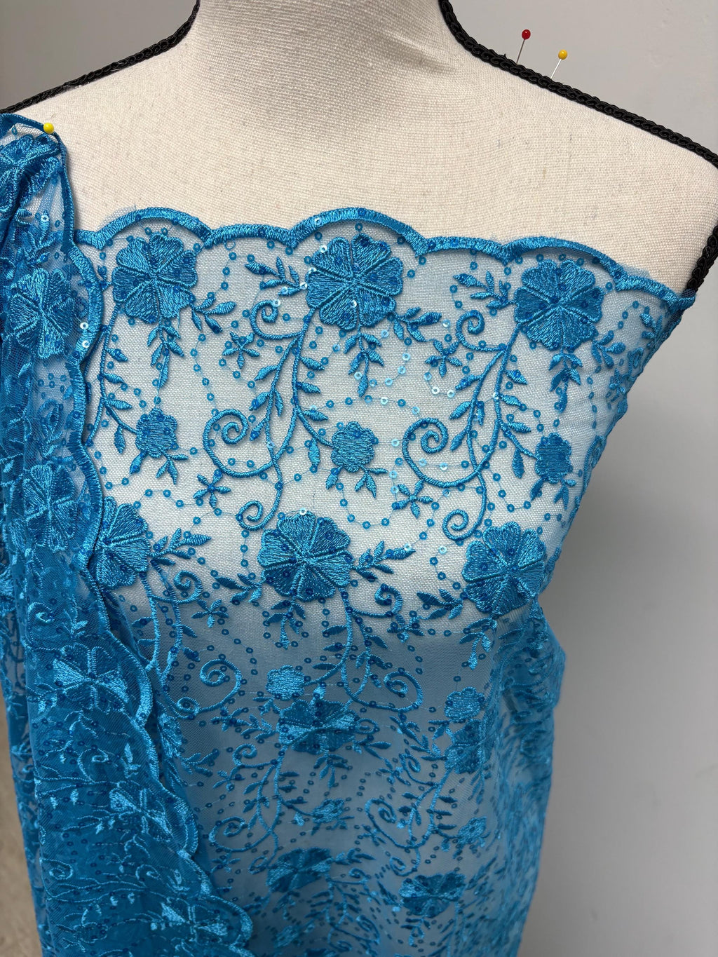 May include: A turquoise lace fabric with an intricate floral design. The fabric features embroidered flowers, leaves, and sequins. The scalloped edge adds a decorative touch. The fabric is draped over a mannequin, showcasing its texture and pattern. This delicate lace is suitable for various sewing and crafting projects.