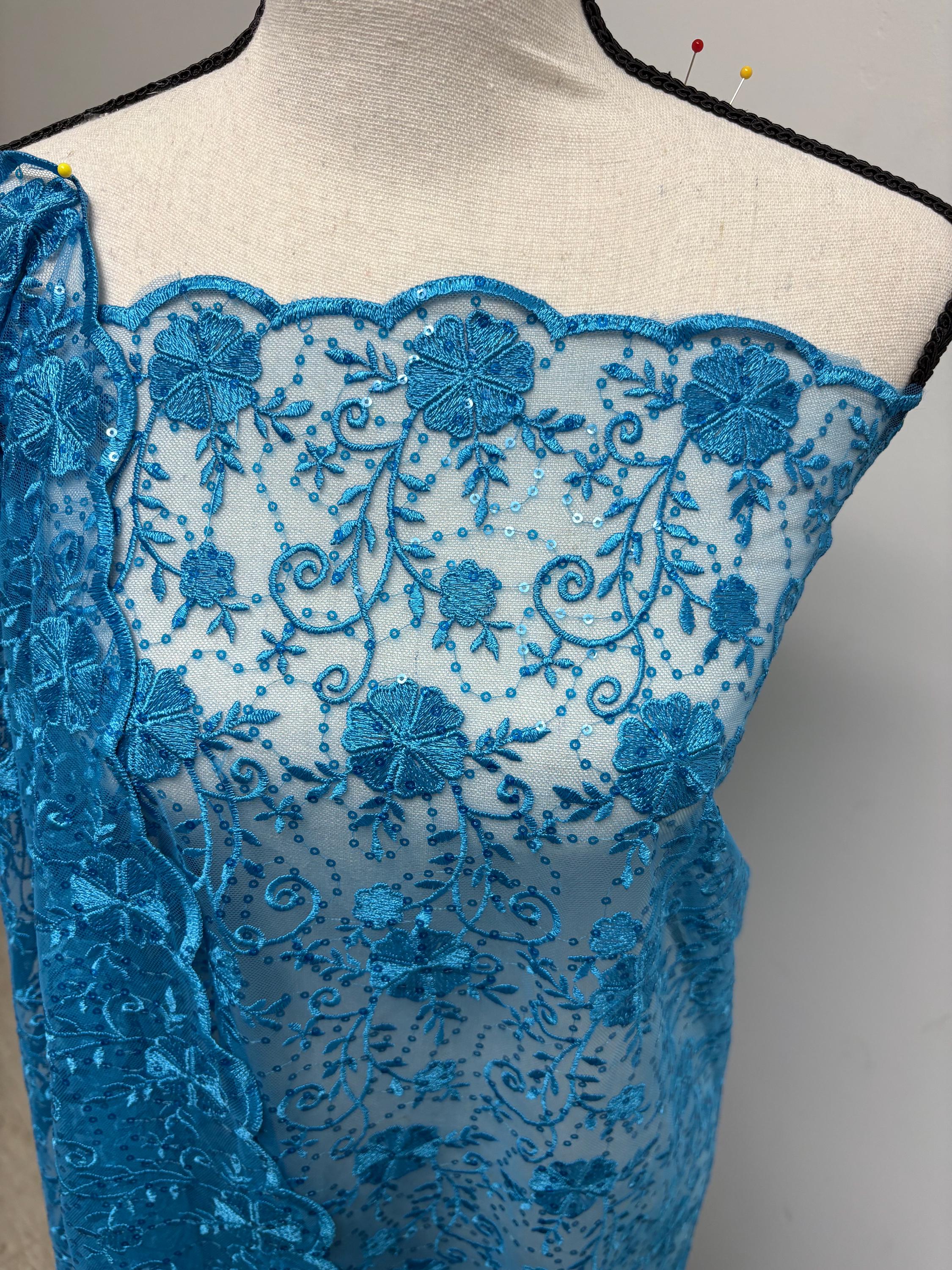 May include: A turquoise lace fabric with an intricate floral design. The fabric features embroidered flowers, leaves, and sequins. The scalloped edge adds a decorative touch. The fabric is draped over a mannequin, showcasing its texture and pattern. This delicate lace is suitable for various sewing and crafting projects.